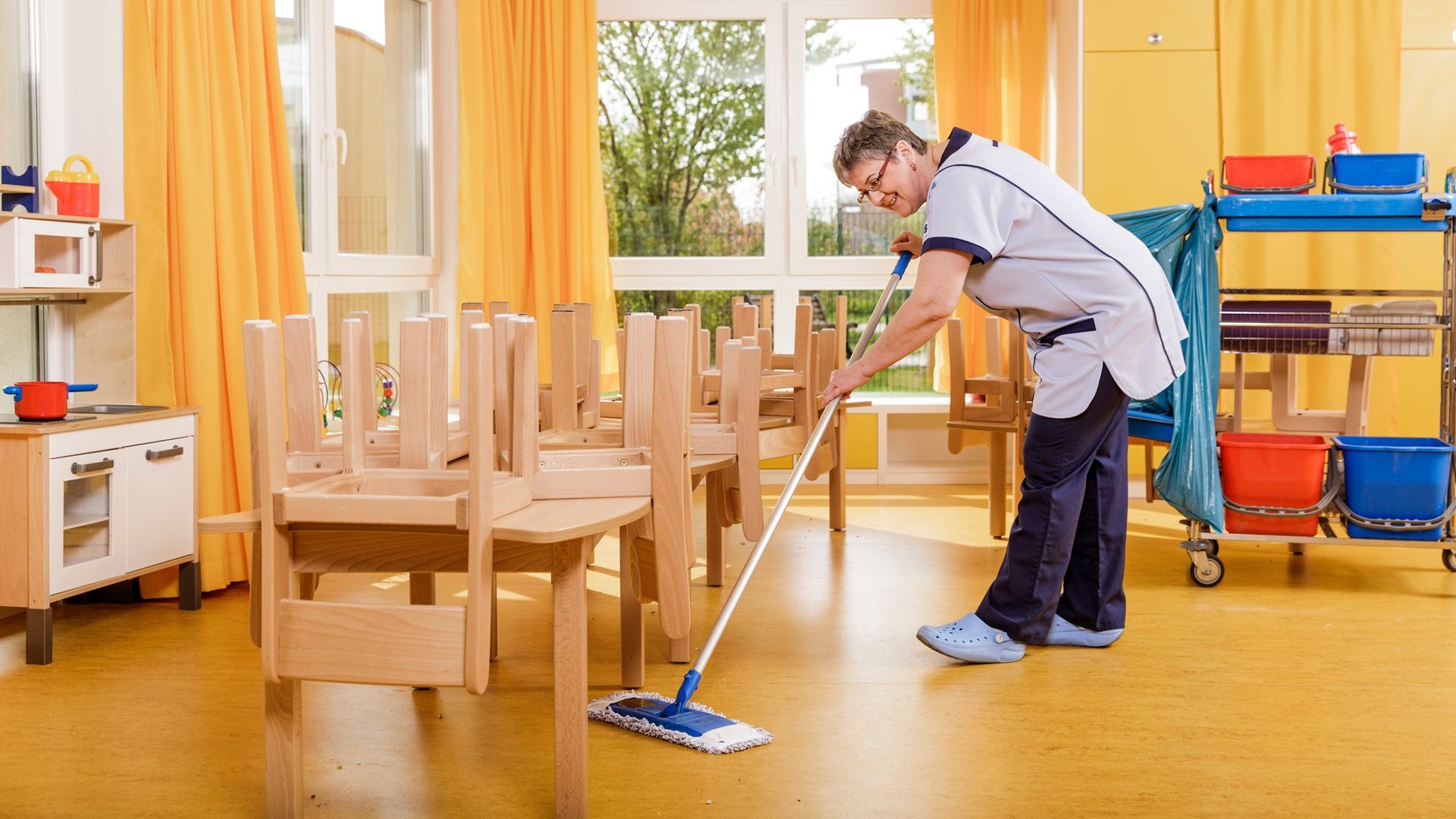 Cleaner mopping the floor of a colorful classroom with chairs stacked on tables and cleaning supplies nearby.