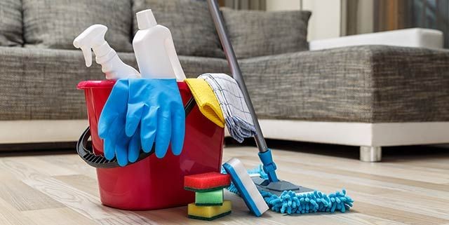 Bucket with cleaning supplies, gloves, and mop on wooden floor near gray sofa.