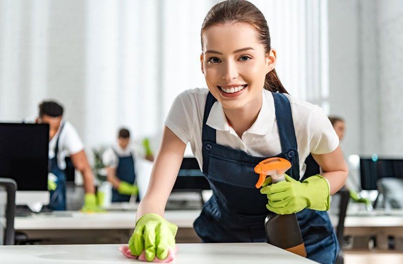 Smiling person wearing gloves, cleaning a desk with spray bottle and cloth in an office setting.