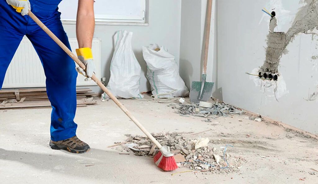 Worker in blue overalls sweeping construction debris with a broom in a partially renovated room.