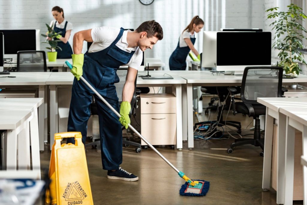 Office cleaning team with mops and gloves working in a modern office space with computers and desks.